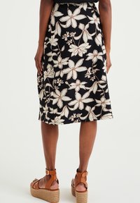 Květinová černá sukně s krémovými a béžovými květinovými vzory, ke kolenům, texturovaná tkanina, v kombinaci s hnědými sandály na klínu se řemínky.
