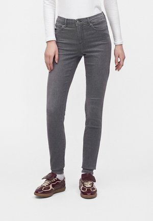 Person iført tætsiddende grå jeans, hvid langærmet skjorte, hvide sokker og mørkerøde sneakers med beige snørebånd, stående mod en hvid baggrund.