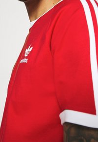 Camiseta atlética roja con acentos y rayas blancas en las mangas, que presenta un pequeño logo blanco de Adidas en el pecho. Textura de tela suave.