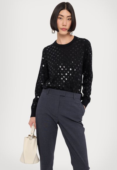 Suéter preto com bolinhas prateadas, mangas longas e decote redondo, combinado com calças cinzas e uma pequena bolsa em creme.