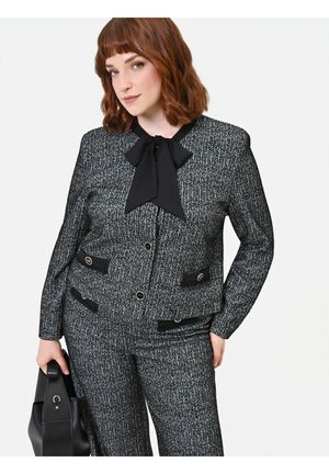 Donna con capelli rossi che indossa un completo bianco e nero con motivo e una camicetta con fiocco nero, che tiene una borsa nera, su sfondo bianco.