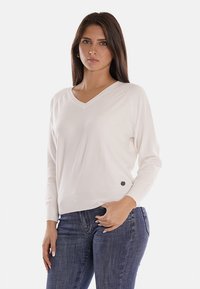 Maglione bianco a V con maniche lunghe, texture liscia e un logo sul lato sinistro. Indossato con jeans blu scuro.