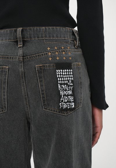 Jeans in denim nero con una vestibilità leggermente ampia. Presentano cuciture incrociate dorate sopra le tasche posteriori e una patch in tessuto nero con testo grafico bianco.
