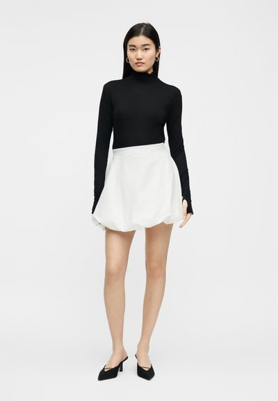 Γυναίκα με μακριά σκουρόχρωμα μαλλιά, ντυμένη με μαύρο μακρυμάνικο ζιβάγκο, λευκή κοντή φούστα και μαύρες μυτερές γόβες, που στέκεται μπροστά σε απλό φόντο.