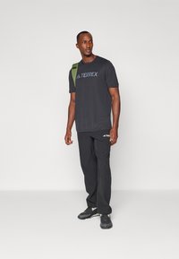 T-shirt de sport noir avec le logo "TERREX", associé à un pantalon noir et des baskets foncées. La personne porte un sac avec une bandoulière verte.