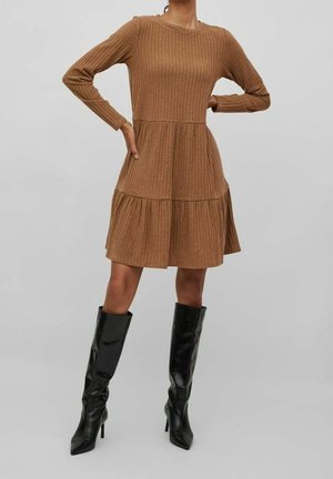 Femme portant une robe marron à côtes avec manches longues et des bottes noires à talons haut montant jusqu’au genou, debout les mains sur les hanches sur un fond uni.