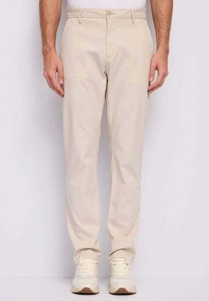 Uomo che indossa pantaloni beige a gamba dritta e sneakers bianche, in piedi contro uno sfondo chiaro uniforme.