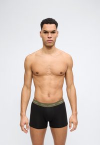 Άνδρας χωρίς πουκάμισο με κοντά σγουρά μαλλιά, φορώντας μαύρα εσώρουχα Tommy Hilfiger με ελαφρώς πράσινη ζώνη μέσης, στέκεται μπροστά σε απλό έντονο φόντο.