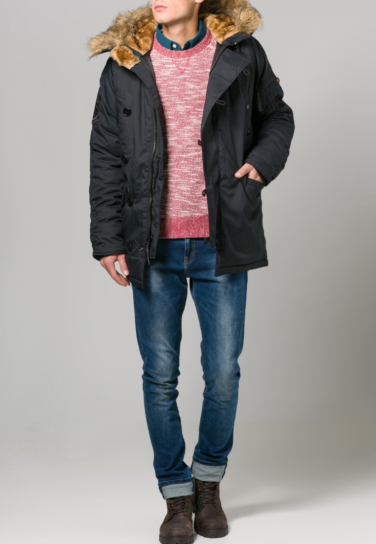 Parka nera imbottita con cappuccio foderato in pelliccia, indossata sopra un maglione rosso con texture, abbinata a jeans blu e stivali marroni con lacci.