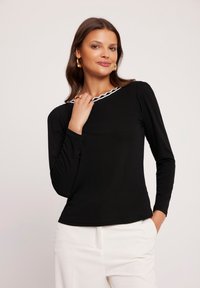 Czarna bluzka z długim rękawem z białą falistą lamówką przy dekolcie, wykonaną z miękkiego materiału, dopasowaną i z lekko bufiastymi rękawami. Zestawiona z białymi spodniami.