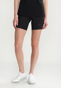 Shorts de sport noirs en tissu extensible, caractérisés par une coupe ajustée. Associés à des baskets blanches avec une semelle transparente.