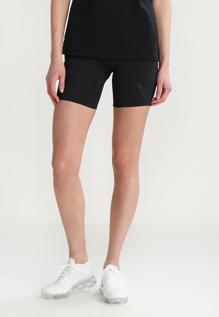 Shorts de sport noirs en tissu extensible, caractérisés par une coupe ajustée. Associés à des baskets blanches avec une semelle transparente.