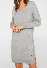 Vestito grigio di maglia con scollo a V, maniche lunghe, spacchi laterali e piccolo dettaglio logo sull'orlo. Tessuto morbido e vestibilità rilassata.