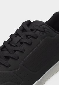 Baskets noires avec des panneaux en cuir texturé, des lacets noirs passés par six œillets, et une semelle en caoutchouc blanche visible sur le bord inférieur.