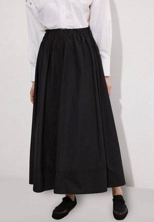 Chemise blanche à manches longues rentrée dans une jupe midi noire taille haute, associée à des baskets noires, debout contre un mur uni.