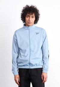Light blue zip-up jacket with a stand-up collar, featuring black side accents and the Reebok logo on the chest. Smooth fabric, elastic cuffs.