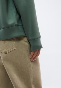 Άτομο που φοράει μπεζ τζιν παντελόνι και πράσινο φούτερ με ριμπ μανσέτες, το χέρι ακουμπάει πλάι στο ισχίο σε απλό ανοιχτό φόντο.