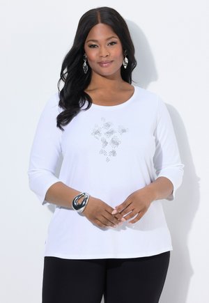 Witte lange mouwen shirt met een ronde hals en zilveren versieringen op de borst, gecombineerd met zwarte broeken en een metalen armband.