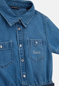 Denim shirt in light blue, featuring a collared neck, snap buttons, two chest pockets, and "Guess" embellished in rhinestones on the left pocket.