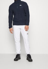 Mörkblå fleecepullover med halvdragkedja, vita byxor och svarta vandringsskor. Pullovern har en liten logotyp och har en mjuk yta.