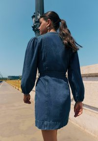 Denimkleit, millel on vormiv vöökoht, puhvitud pikad varrukad ja põlve keskpaigani ulatuv pikkus. Tume sinine kangas, sileda tekstuuri ja peente õmblustega.