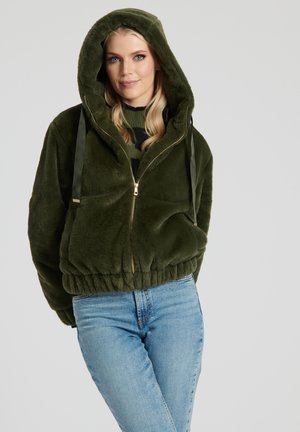 Hoodie en peluche vert kaki avec fermeture éclair, cordons de serrage, ourlet élastique et accents dorés. Associé à un jean en denim bleu clair.