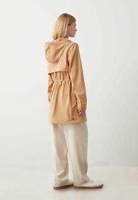 Helle beige wasserfeste Jacke mit einer Kordelzugtaille, Kapuze und kontrastierenden Hardwaredetails, kombiniert mit cremefarbenen, weit geschnittenen Hosen und weißen Sneakern.