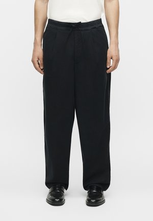 Άνδρας που φοράει μαύρο παντελόνι με κορδόνι, μαύρα επίσημα παπούτσια και λευκό κοντομάνικο πουκάμισο σε απλό φόντο.