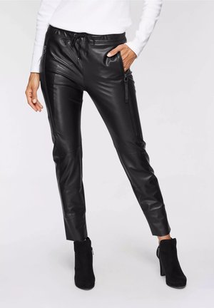 Donna che indossa pantaloni neri in ecopelle con cerniere frontali e stivaletti neri, mano in tasca su sfondo neutro.