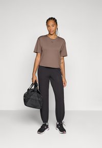 Brun T-skjorte med kort erm, sorte joggebukser og svarte joggesko; holder en svart treningsbag med nett detaljer. Minimalistisk treningstøy.