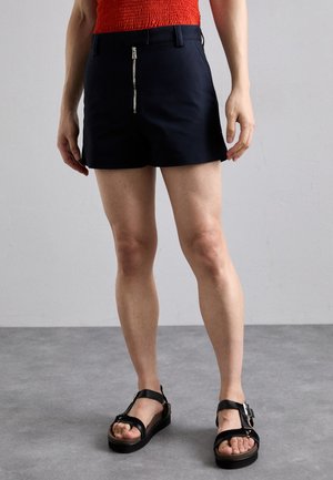 Marineblaue Shorts mit einem Frontreißverschluss, flachem Design und Gürtelschlaufen. Getragen mit schwarzen Sandalen mit Riemen auf einer hellgrauen Oberfläche.