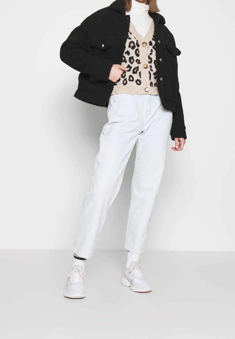 Zwarte fleecejas met kraag, beige vest met luipaardpatroon, lichtblauwe taps toelopende spijkerbroek en witte sneakers. Textuur van de stof en knopen zichtbaar.