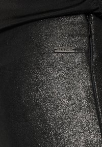 Zwarte leggings met een glinsterende textuur, voorzien van een klein metalen logo-label. De stof heeft een glanzende, reflecterende kwaliteit.