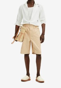 Pantalones cortos de algodón beige con dos bolsillos frontales, combinados con una camisa blanca de manga larga con detalles bordados en negro. Bolsa de paja trenzada y zapatos claros.