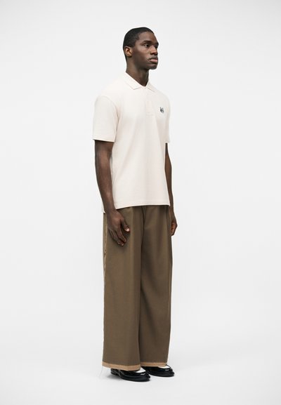 Άνδρας που στέκεται φορώντας άσπρο πόλο μπλουζάκι με κοντά μανίκια και μικρό λογότυπο στο στήθος, φαρδιά καφέ παντελόνια και μαύρα παπούτσια, σε λευκό φόντο.