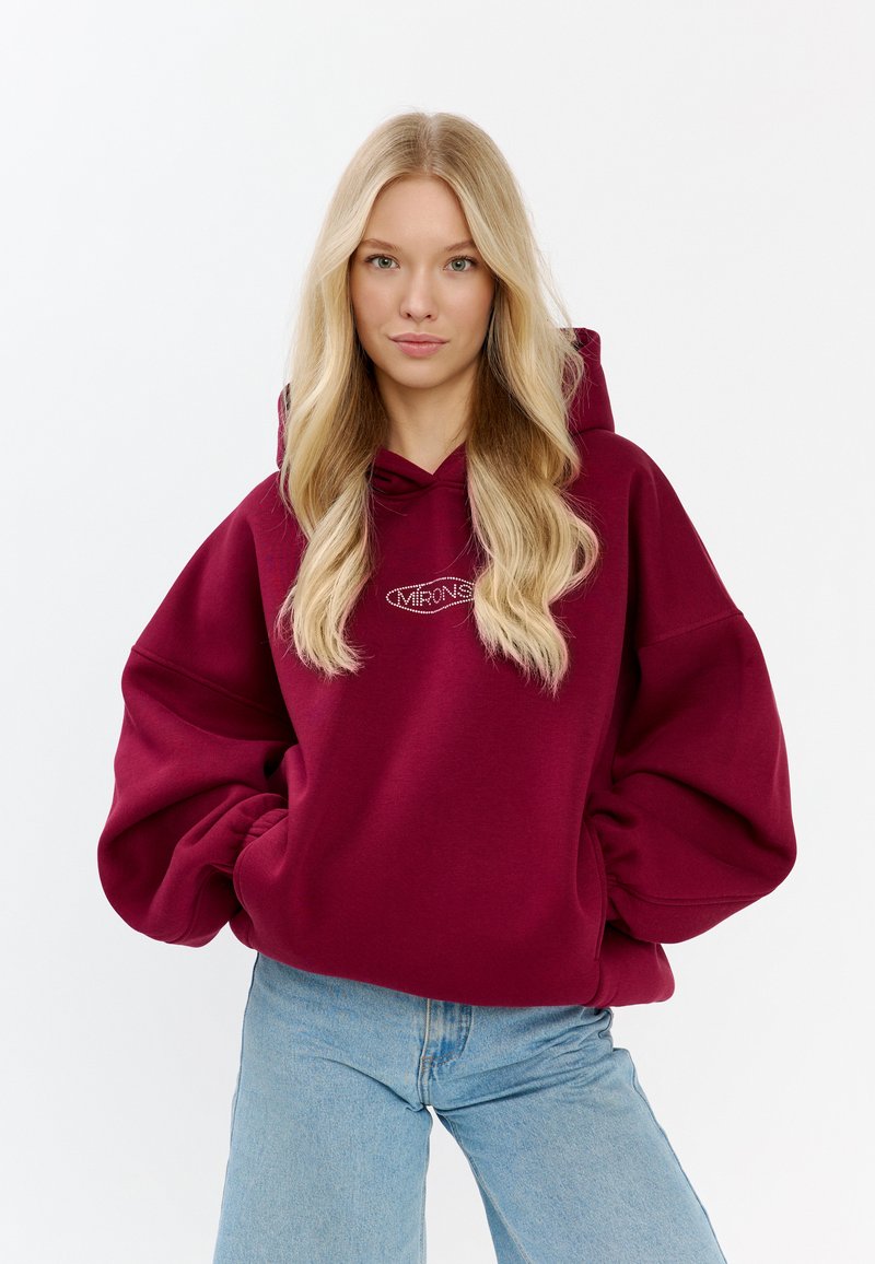 Hoodie oversize color borgogna con cappuccio, polsini a costine e logo ricamato sul petto. Abbinato a jeans azzurri chiari.