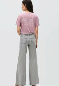 Růžové bavlněné tričko s bílým textem na zádech, kombinováno s šedými džínami s vysokým pasem a širokými nohavicemi. Hladká textura, volný střih.