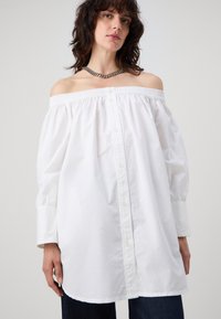Camisetta bianca off-the-shoulder con bordi elasticizzati, apertura a bottoni frontale e maniche lunghe a palloncino. Realizzata in tessuto leggero, leggermente testurizzato.