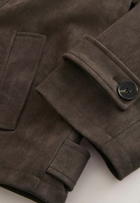 Ruda netikro zomšos striukė su struktūruotu apykakliu, atvartų kišenėmis ir sagos detalėmis rankogalyje, lygios tekstūros.