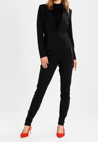 Blazer negro ajustado con un botón, acompañado de pantalones negros ajustados. Presenta tacones rojos. Textura suave en toda la prenda con un diseño estructurado.