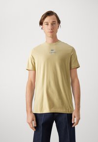 Svietložlté bavlnené tričko s krátkymi rukávmi a okrúhlym výstrihom, na prednej strane s umiestneným zeleným logom Lacoste a textom "LACOSTE".