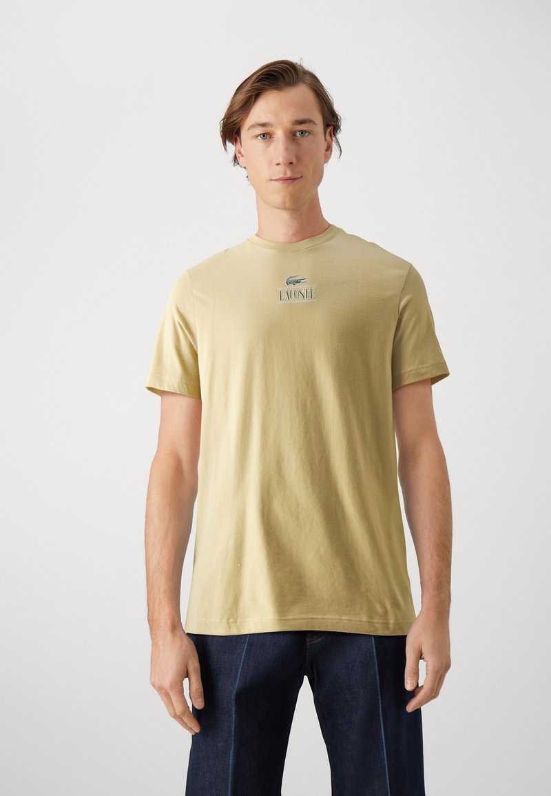 Svietložlté bavlnené tričko s krátkymi rukávmi a okrúhlym výstrihom, na prednej strane s umiestneným zeleným logom Lacoste a textom "LACOSTE".
