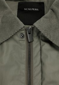 Oliven grøn jakke med fløjlskrave, synligt Scalpers-mærke og metal lynlåstrækker med logo.
