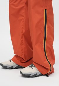 Oranžas ūdensizturīgas bikses ar sānu rāvējslēdzēju, rotātas ar ziliem un dzelteniem svītrām, saskaņotas ar pelēkām un bēšām sporta kurpēm ar teksturētām zolēm.