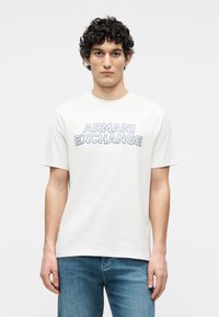 Νεαρός άνδρας με σγουρά μαλλιά που φοράει λευκό T-shirt Armani Exchange και μπλε τζιν, στέκεται μπροστά σε απλό ανοιχτό φόντο.