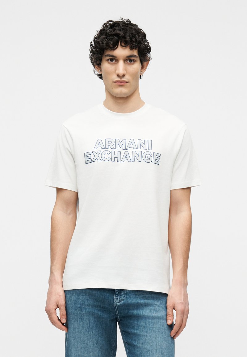 Ν εαρός άνδρας με σγουρά μαλλιά που φοράει λευκό T-shirt Armani Exchange και μπλε τζιν, στέκεται μπροστά σε απλό ανοιχτό φόντο.