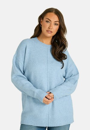 Femme aux longs cheveux foncés porte un pull bleu clair ample et un jean bleu, debout, les mains jointes devant un fond uni.
