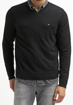Mann trägt einen schwarzen Pullover mit V-Ausschnitt über einem grauen Hemd mit Knöpfen und hellgrauen Jeans, steht vor weißem Hintergrund.