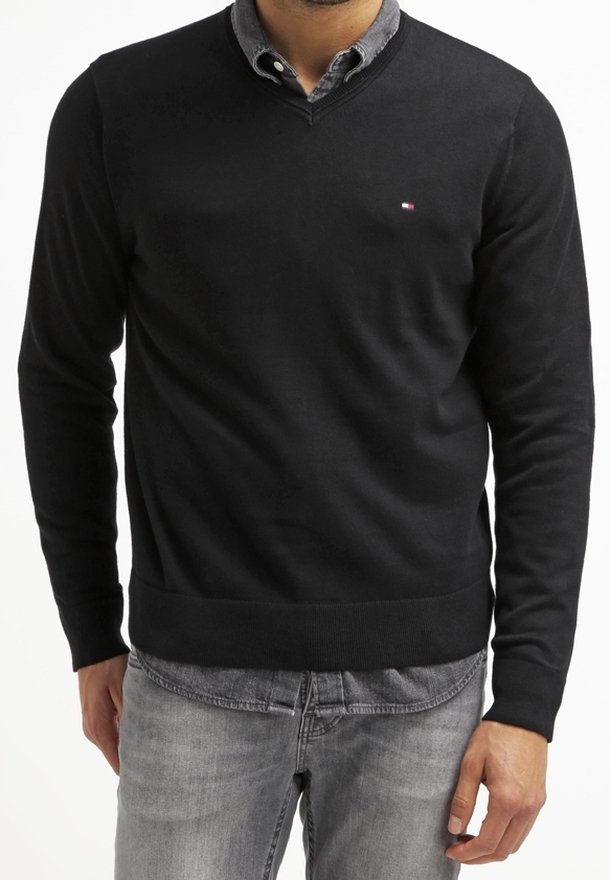 Homme portant un pull noir à col en V par-dessus une chemise grise à boutons et un jean gris clair, debout devant un fond blanc.