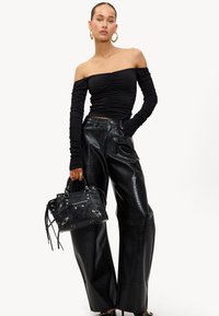 Vrouw in een zwarte gerimpelde off-shoulder top, zwarte wijdere nepleren broek, met een zwarte gestructureerde handtas, poserend voor een witte achtergrond.